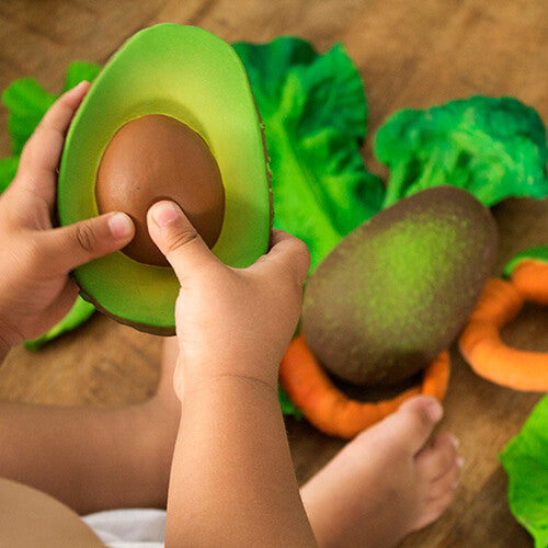Kinderhandjes houden het oli & carol bijtspeelgoed in de vorm van een avocado vast, met ander groentespeelgoed op de achtergrond.