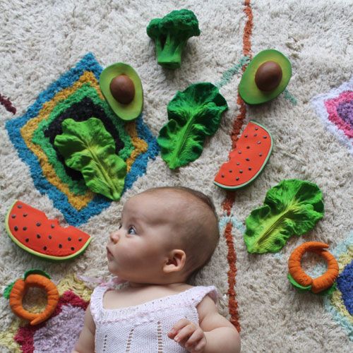 Een baby ligt op een kleed omringd door oli & carol bijtspeelgoed in de vorm van fruit en groenten.