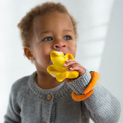 Een baby bijt op een Oli & Carol banaan bijtspeeltje en draagt een wortel bijtspeeltje om de pols.