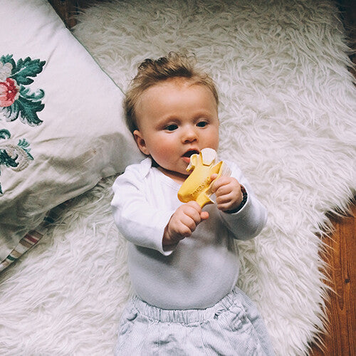 Een baby ligt op een witte vacht en houdt een Oli & Carol banaan bijtspeeltje vast.