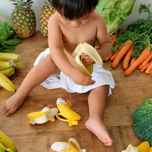 Een baby speelt op de grond met een Oli & Carol banaan bijtspeeltje, omringd door echt fruit en groenten.