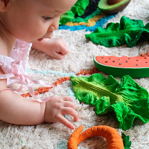 Een baby speelt op een kleed met verschillende Oli & Carol groenten en fruit bijtspeeltjes.