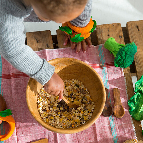 Een kind speelt met een kom granen, met het groene broccoli speelgoed en andere groenten speelgoed op de achtergrond.
