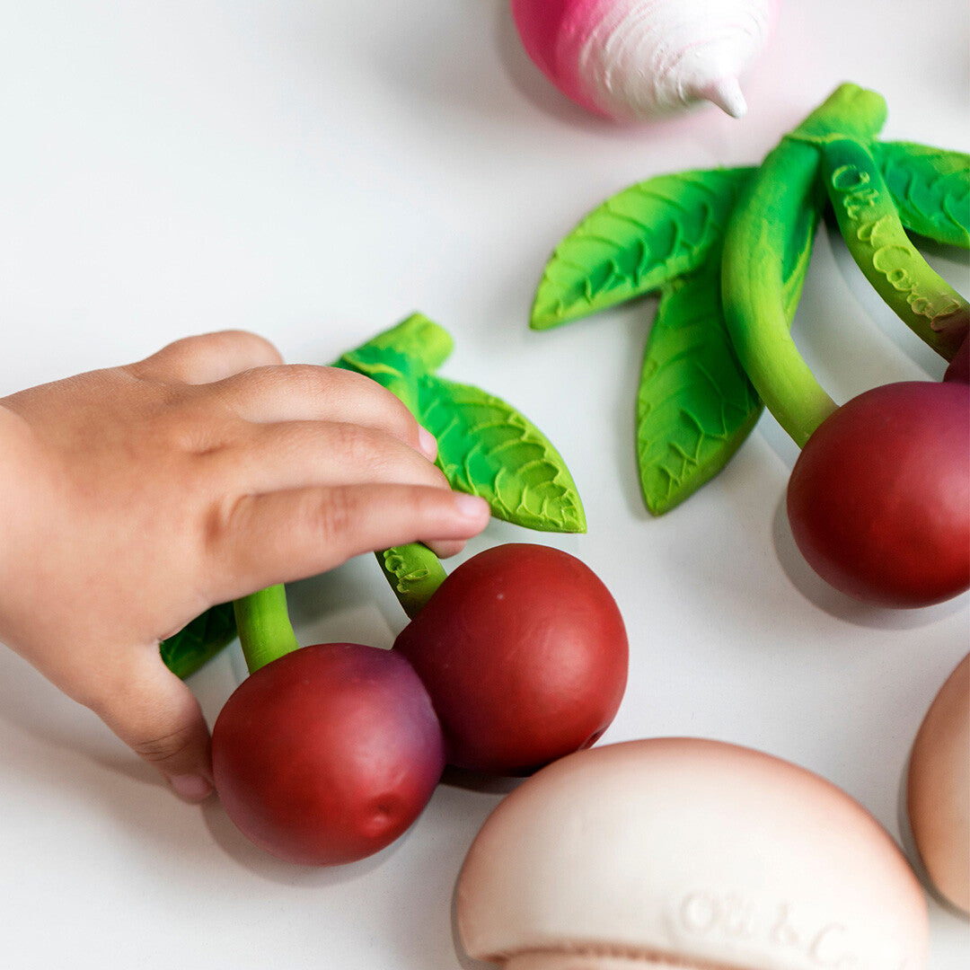 Een kinderhand pakt een rood kersen bijtspeeltje van rubber, liggend tussen ander fruit- en groentespeelgoed.