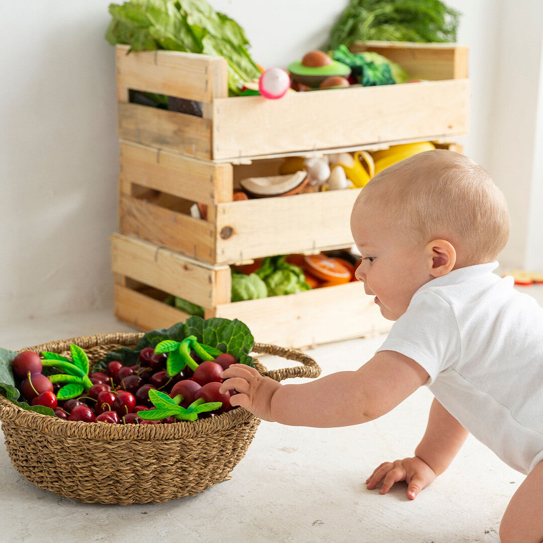 Een baby kruipt naar een mand vol met Oli & Carol kersen bijtspeeltjes en ander fruit- en groentespeelgoed.