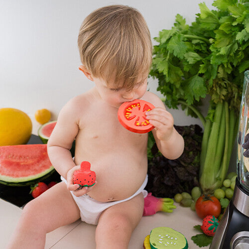 Een baby bijt op het rode tomaten bijtspeelgoed, omringd door ander speelgoed en echt fruit.