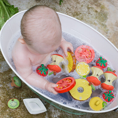 Een baby speelt in bad met diverse fruitvormige badspeeltjes, waaronder het rode tomaten badspeelgoed.