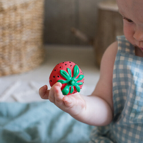 Een babyhandje houdt het Oli & Carol bijtspeeltje in de vorm van een rode aardbei vast.