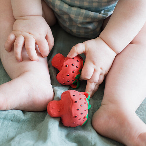 Twee Oli & Carol bijtspeeltjes in de vorm van rode aardbeien liggen op een groene deken, met babybeentjes en -handjes erbij.