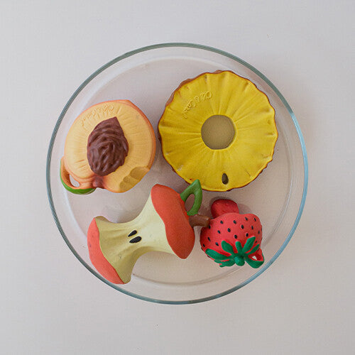 Een collectie Oli & Carol bijtspeeltjes in de vorm van fruit, waaronder een aardbei, ananas, perzik en appelklokhuis, op een glazen schaal.