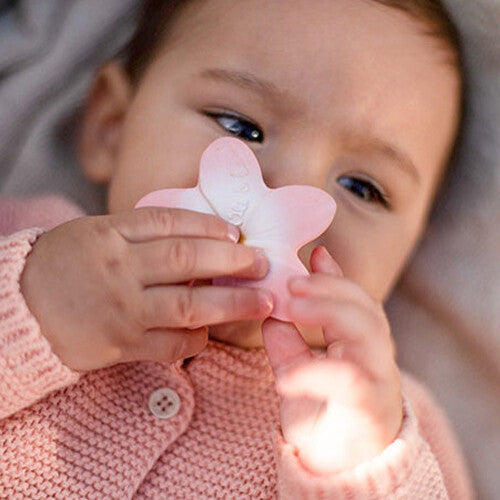 Een baby met een roze gebreid vestje houdt het Oli & Carol babyspeelgoed bijtring in de vorm van een roze plumeria bloem tegen de mond.