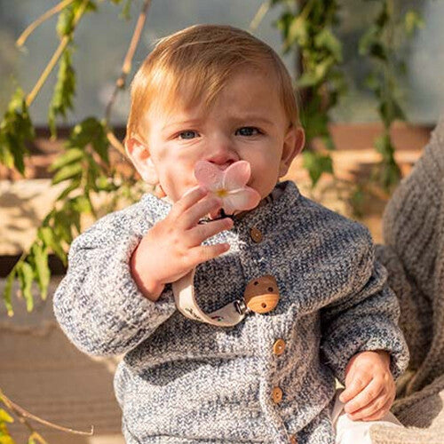 Een baby met een grijs gebreid vestje houdt het Oli & Carol babyspeelgoed bijtring in de vorm van een roze plumeria bloem tegen de neus.