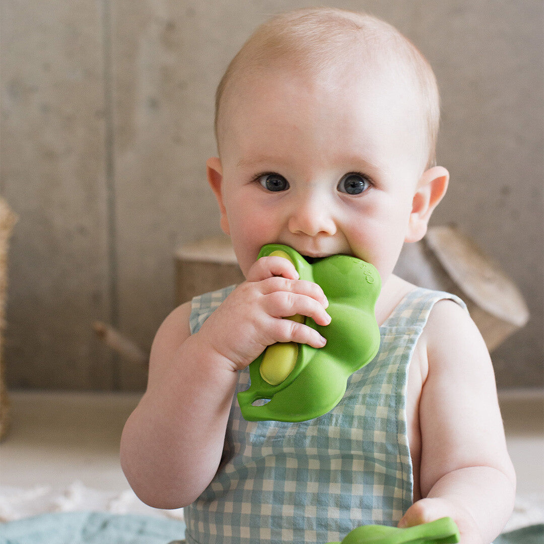 Een baby met een geruite romper bijt op het groene edamame bijtspeeltje.