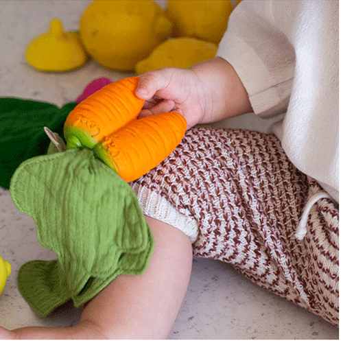 Een babyhandje houdt het Oli & Carol wortel bijtspeeltje met groene stoffen bladeren vast.