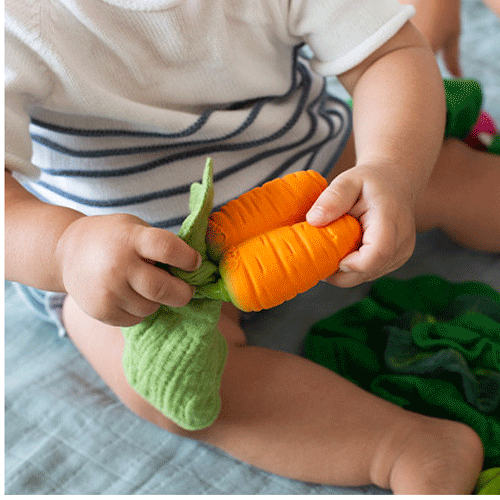 Een baby houdt het Oli & Carol wortel bijtspeeltje met groene stoffen bladeren vast met beide handen.