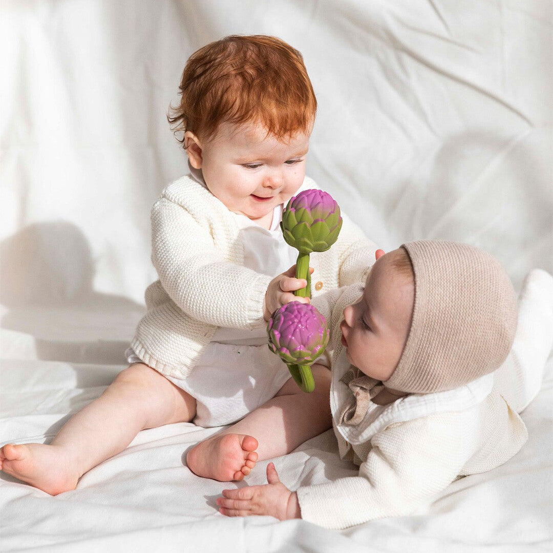 Twee baby's spelen samen met de Oli & Carol artisjok rammelaars op een witte deken.