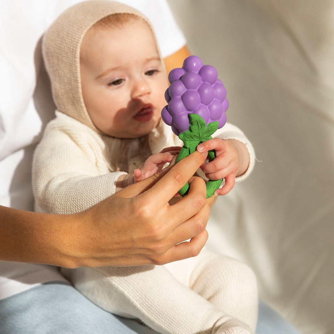 Een baby in een lichte outfit houdt de Oli & Carol druiven rammelaar vast, geholpen door volwassen handen.