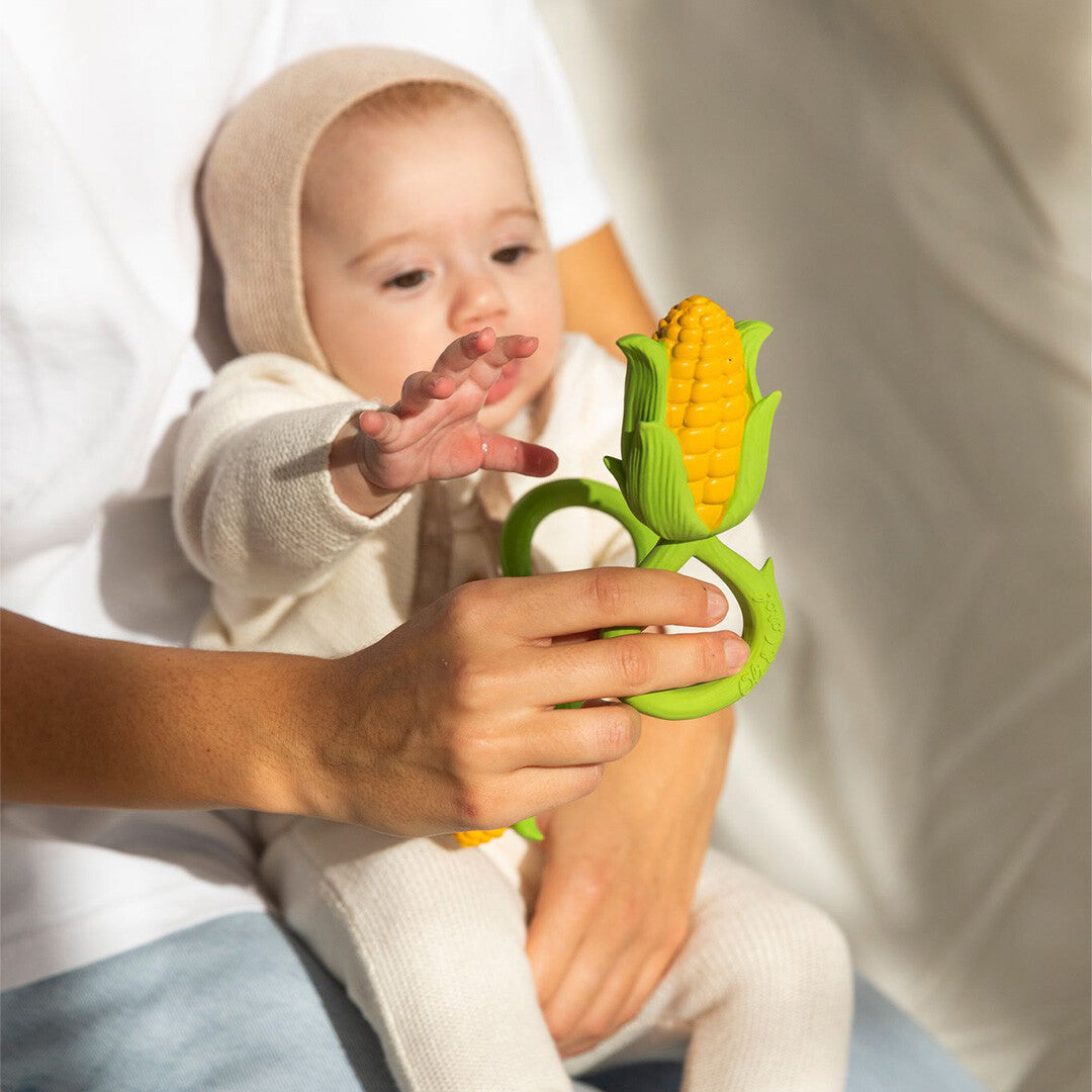 Een baby kijkt naar het Oli & Carol maïs bijtspeelgoed dat een volwassene vasthoudt.