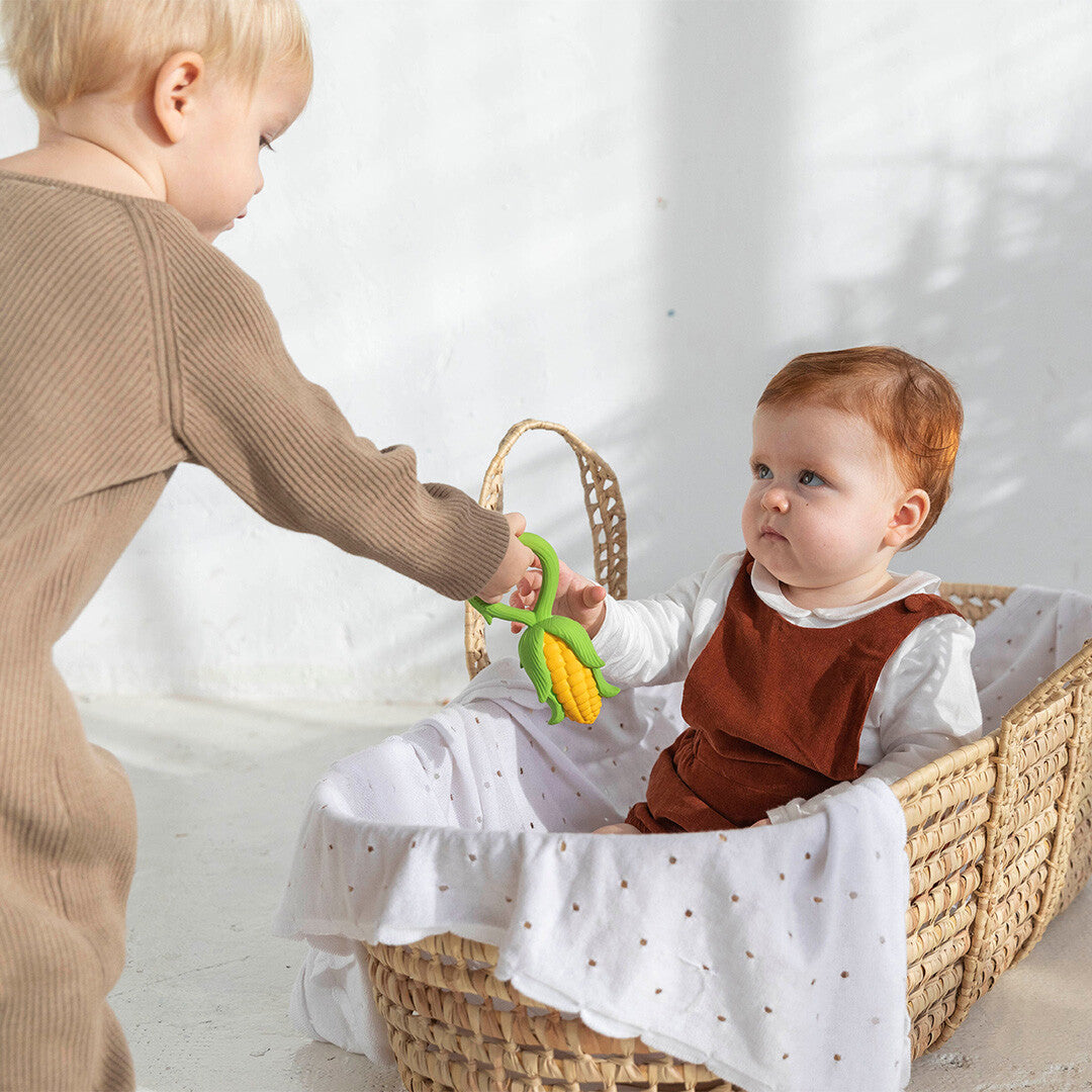 Een ouder kind geeft het Oli & Carol maïs bijtspeelgoed aan een baby in een rieten mand.