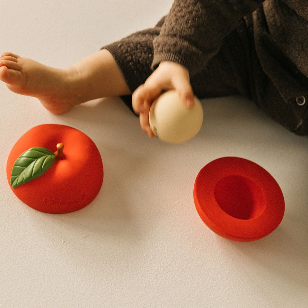 Een baby speelt met de Oli & Carol 3-in-1 babyspeelbal, waarbij de crèmekleurige rammelaar uit de rode appelhelften is gehaald.