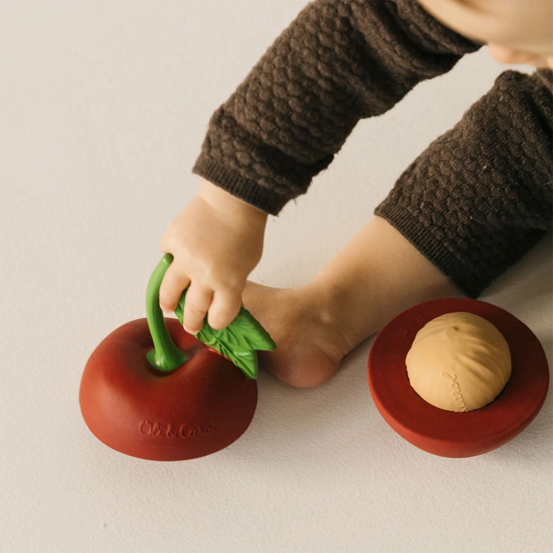 Een babyhandje pakt de Oli & Carol 3-in-1 babyspeelbal kers, die opengeklapt op de grond ligt.