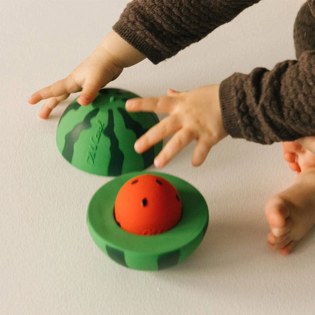 Babyhandjes spelen met het Oli & Carol watermeloen babyspeelgoed dat open op de grond ligt.