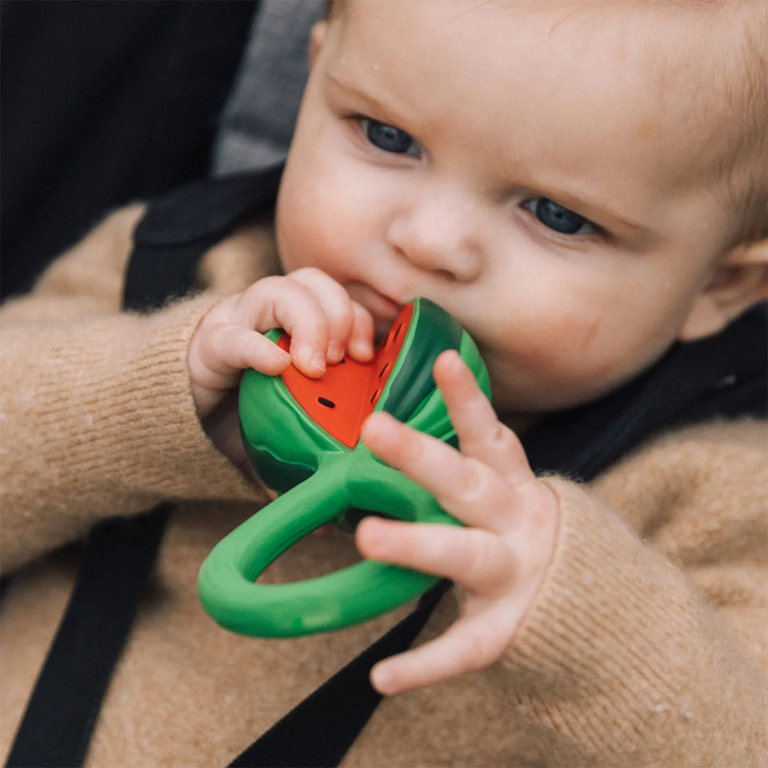 Een baby met blauwe ogen kauwt op de groene Oli & Carol watermeloen rammelaar.