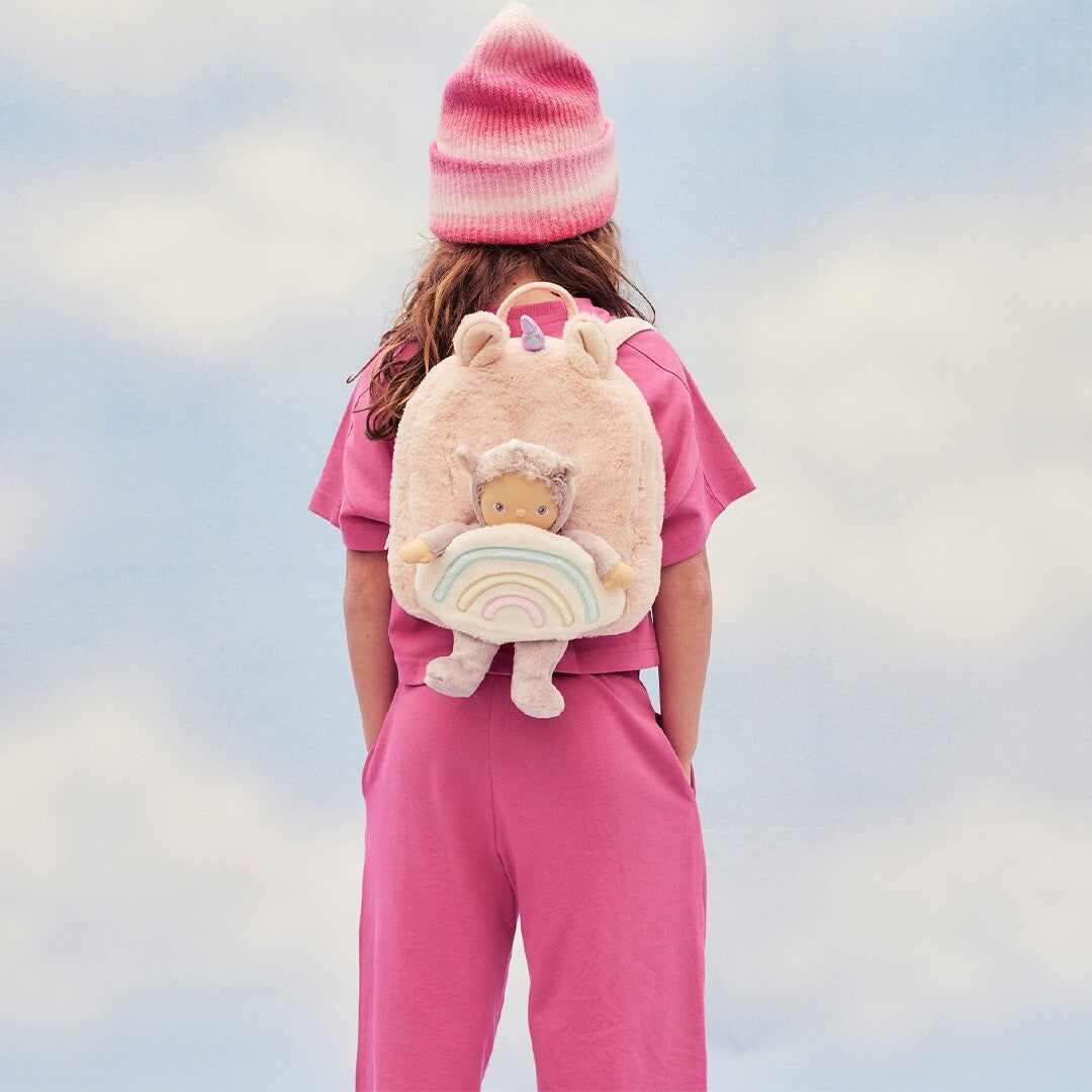Kind met roze kleding en muts draagt de Olli Ella roze eenhoorn kleuterrugzak met pop op de rug.