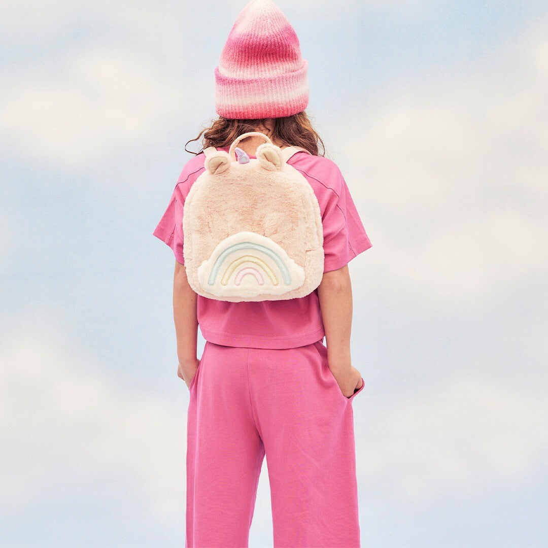 Kind met roze kleding en muts draagt de Olli Ella roze eenhoorn kleuterrugzak met regenboog op de rug.