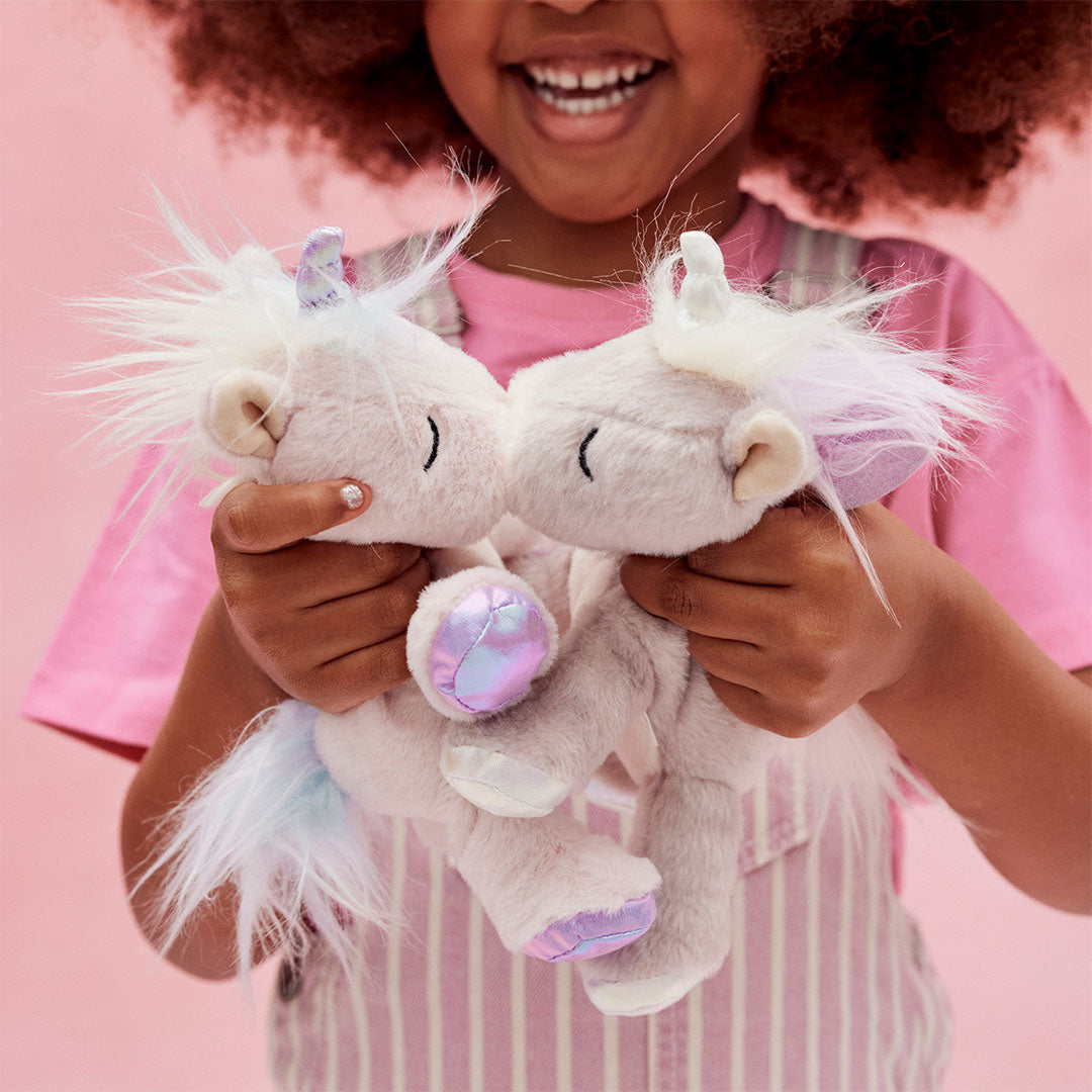 Twee olli ella knuffeleenhoorns Binky Baby Unicorns die elkaar kusjes geven, vastgehouden door kinderhanden.