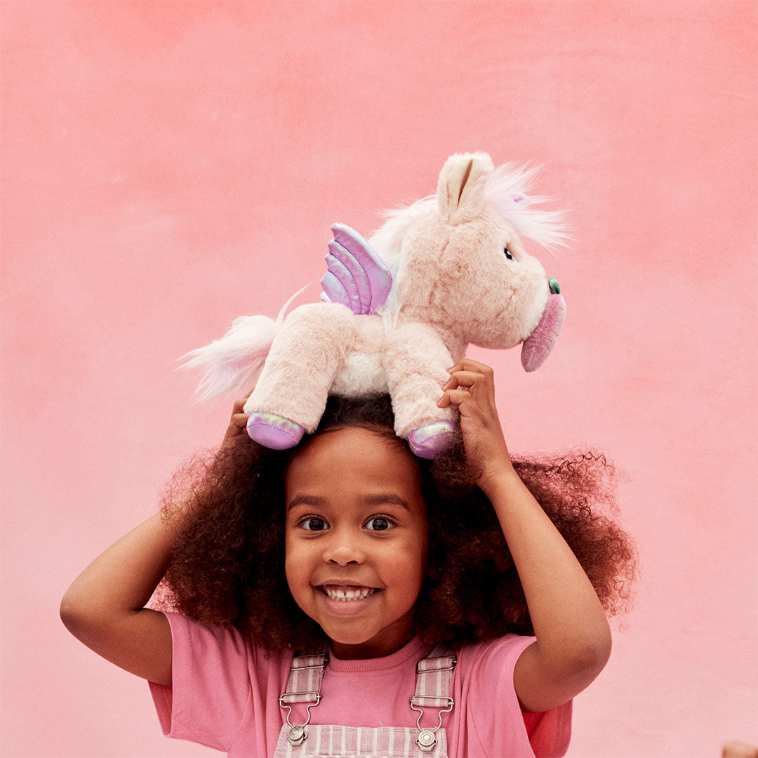Een lachend kind met krullend haar houdt de roze olli ella pluchen eenhoorn knuffel op haar hoofd.