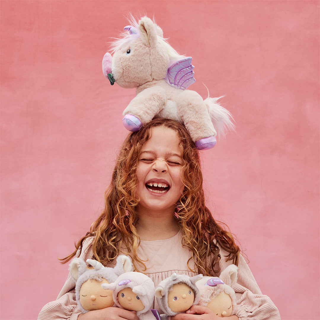 Een lachend kind met de roze olli ella pluchen eenhoorn knuffel op haar hoofd en drie andere knuffelpoppen in haar armen.