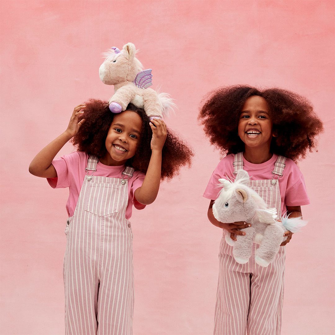 Twee kinderen met krullend haar, één met de roze olli ella pluchen eenhoorn knuffel op haar hoofd en de ander met een grijze eenhoorn knuffel.
