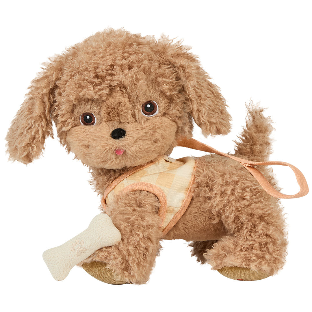 Olli Ella knuffelhond Goldie, een bruine pluchen hond met geruit tuigje en een botje in de bek, staat schuin.