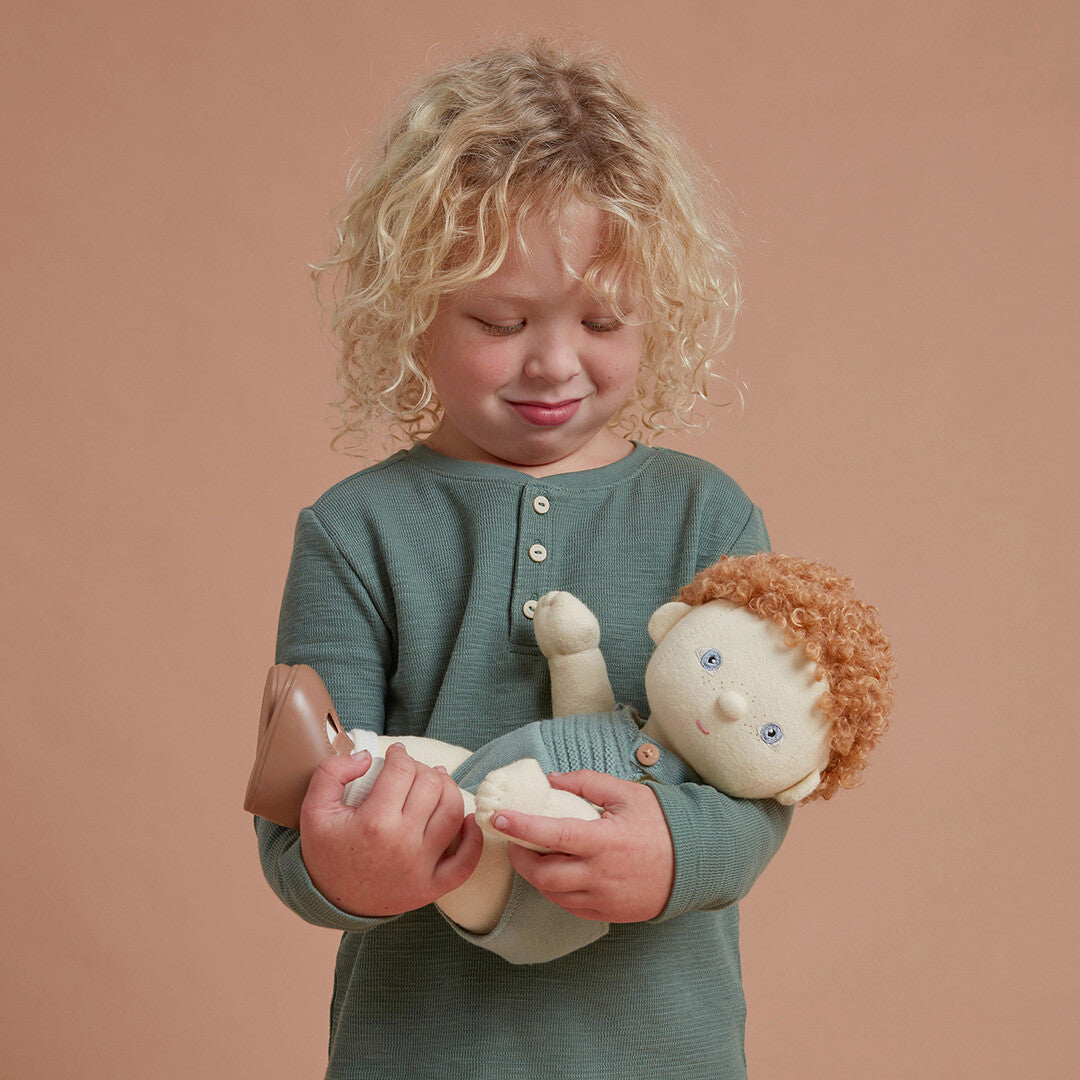 Een kind met blond krullend haar houdt de Olli Ella lappenpop Dinkum Doll met rood haar en groene romper vast.