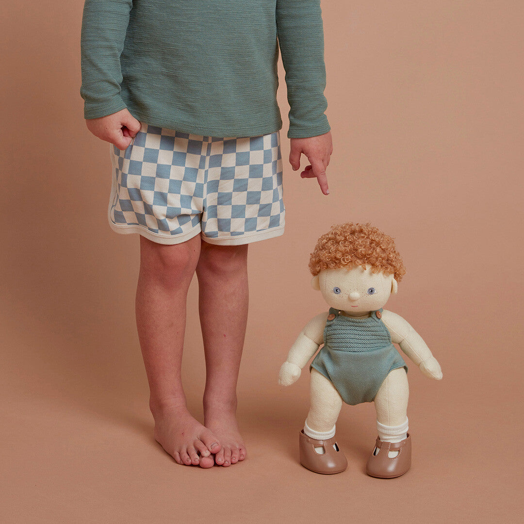 De Olli Ella lappenpop Dinkum Doll met rood haar en groene romper staat naast de benen van een kind.