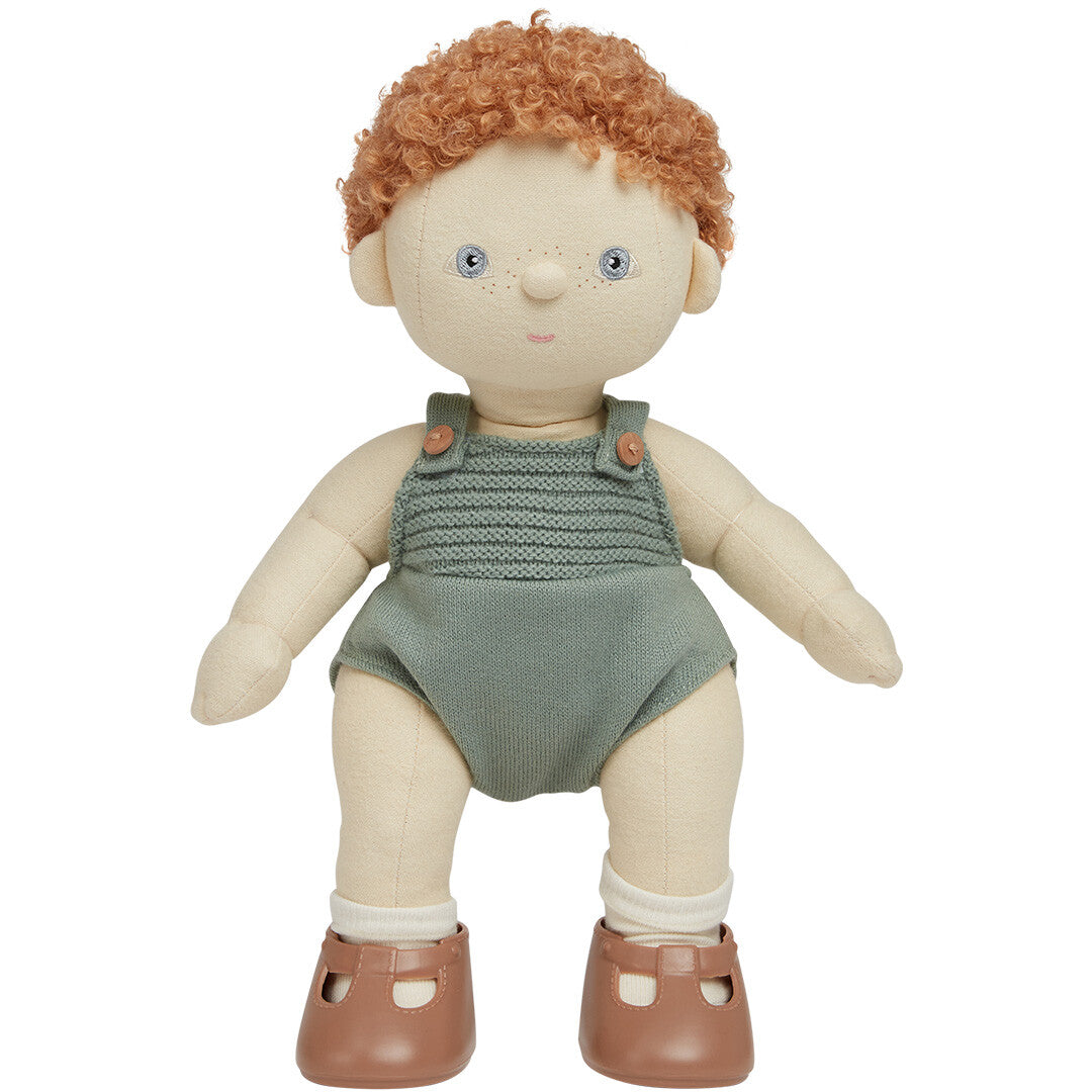 Olli Ella lappenpop Dinkum Doll met rood krullend haar, staand in een groene gebreide romper met witte sokken en bruine schoenen.