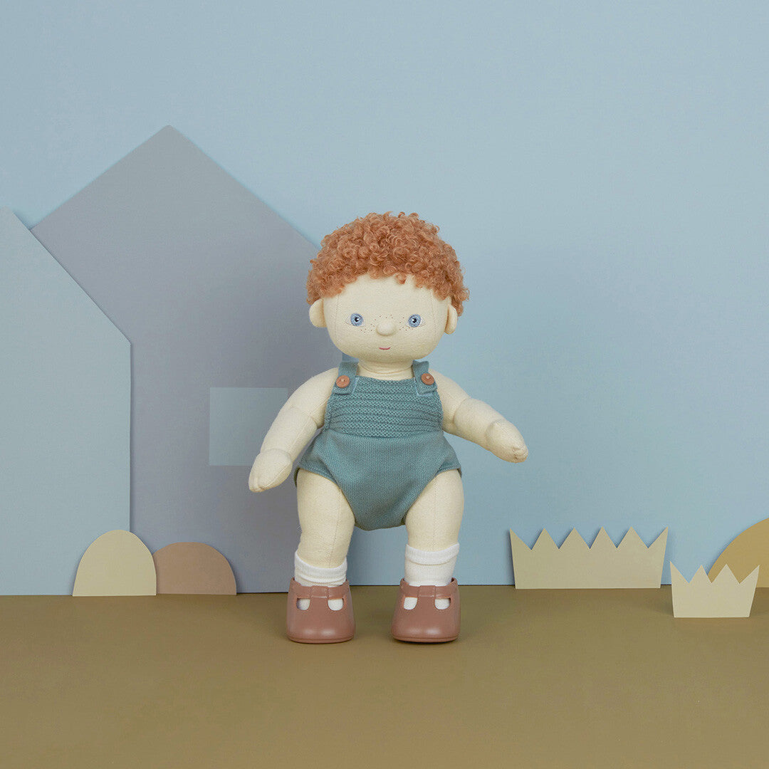 Olli Ella lappenpop Dinkum Doll met rood krullend haar, staand in een groene gebreide romper in een speelse setting.