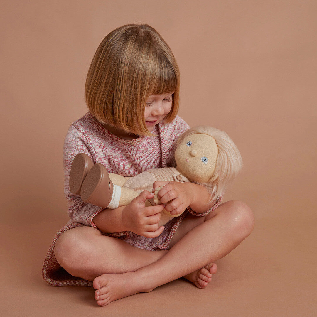 Een meisje met blond haar zit met gekruiste benen en houdt de Olli Ella lappenpop liefdevol vast.