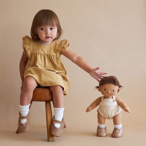Een jong kind zit op een krukje en reikt naar een staande Olli Ella lappenpop Dinkum Doll.