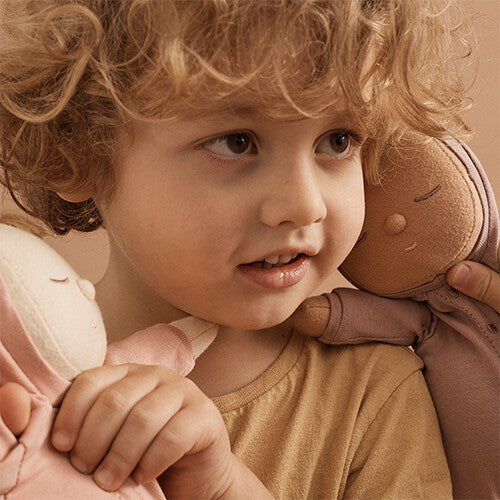 Een kind met krullen houdt twee Olli Ella lappenpoppen vast, een roze en een bruine.