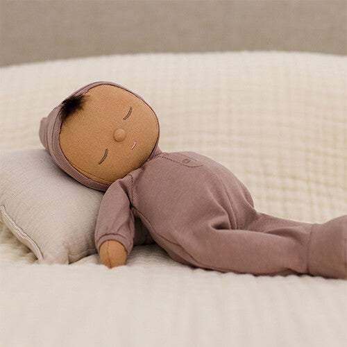 Olli Ella lappenpop Dozy Dinkum Doll Pip in roze onesie met regenboogje, liggend op een bed met kussen.