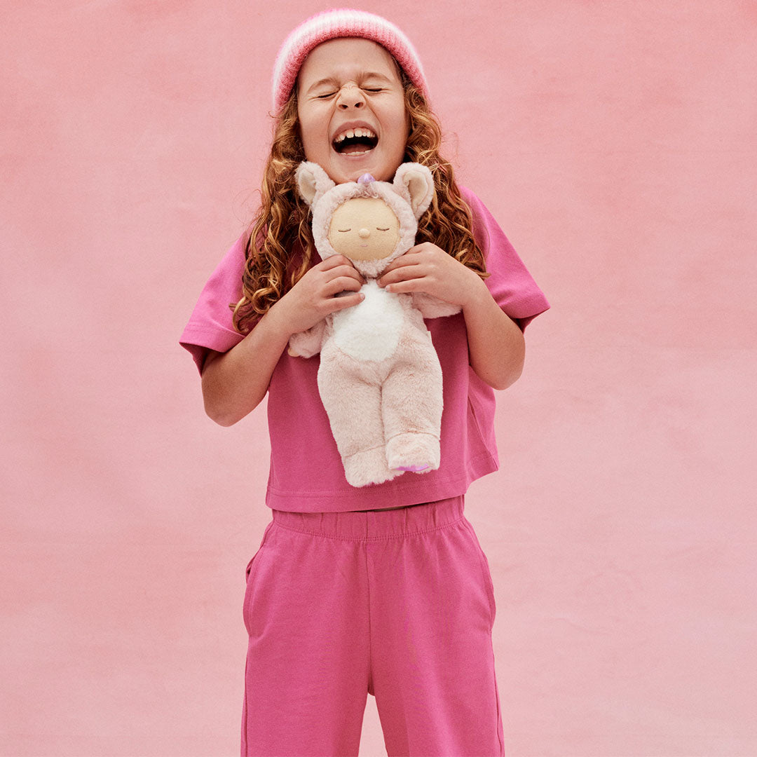 Een lachend kind in roze kleding en gestreepte muts houdt de Olli Ella roze pluche eenhoorn lappenpop vast.