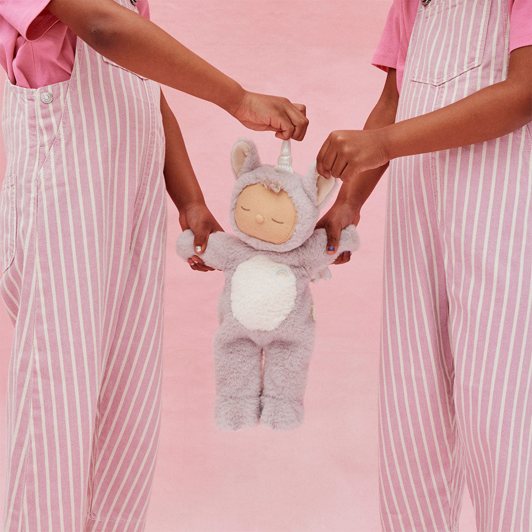 Twee kinderen in roze gestreepte overalls houden samen de olli ella lappenpop eenhoorn Cozy Dinkum Doll Sweetpea vast tegen een roze achtergrond.