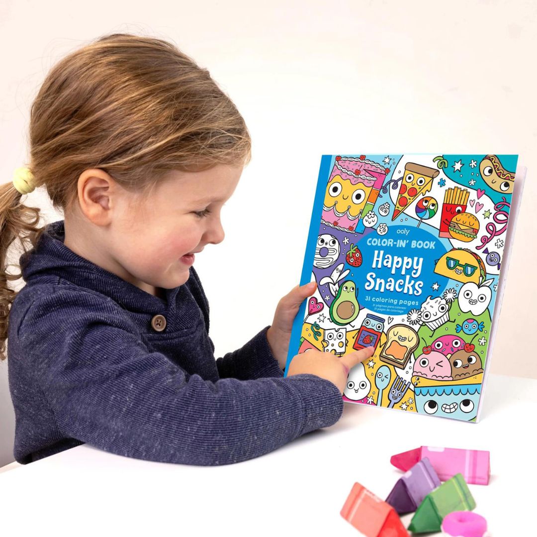 Een kind wijst naar de voorkant van het ooly kleurboek 'Happy Snacks' aan een witte tafel.