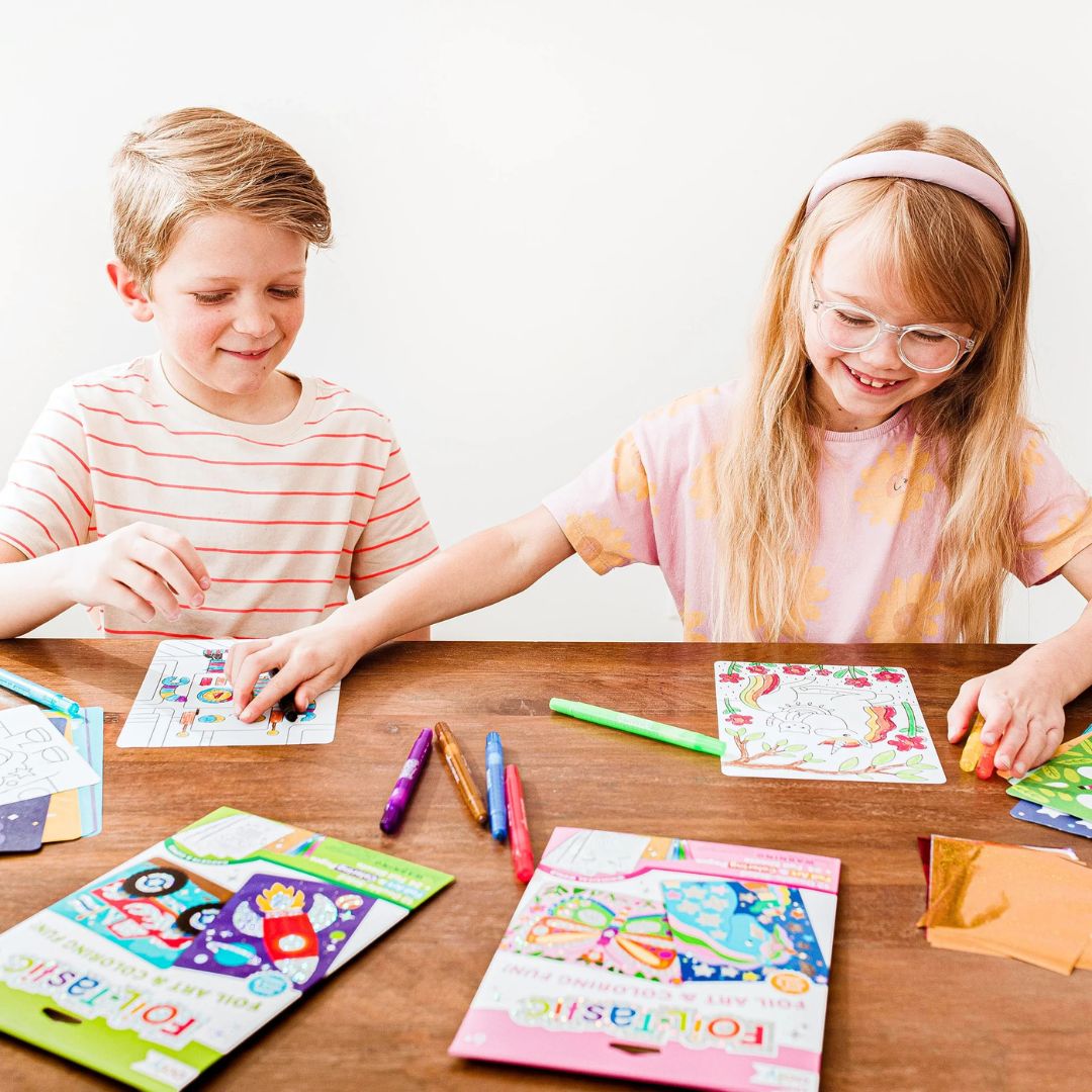 Twee kinderen maken vrolijk folie kunst met de ooly Foil-Tastic knutselset aan een houten tafel.
