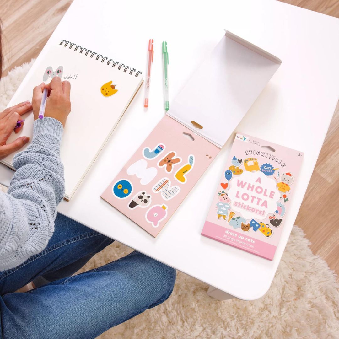 Een persoon plakt ooly stickers uit een stickerboek in een notitieboekje op een witte tafel.