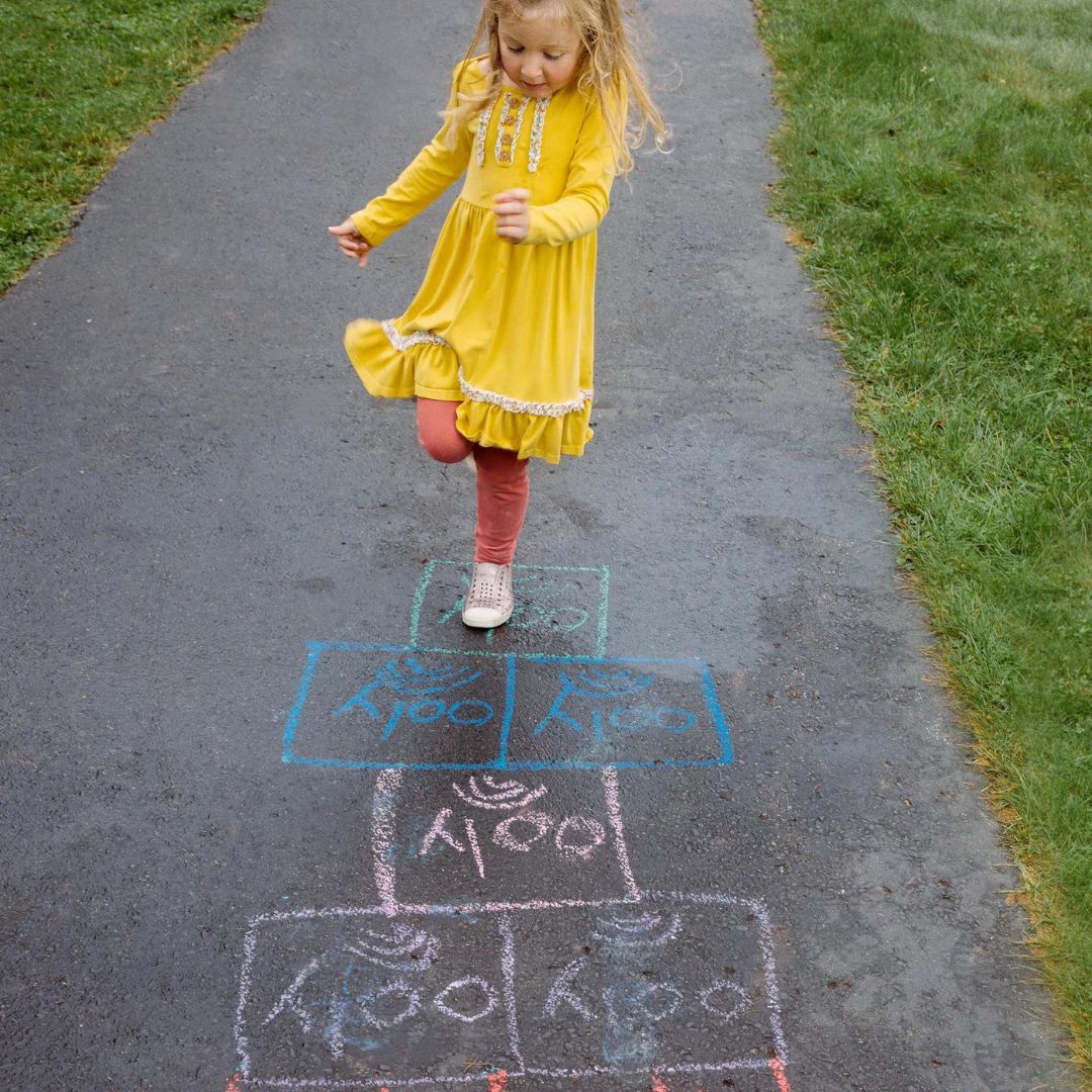 Een kind in een gele jurk speelt hinkelspel op een pad, getekend met kleurrijk stoepkrijt.