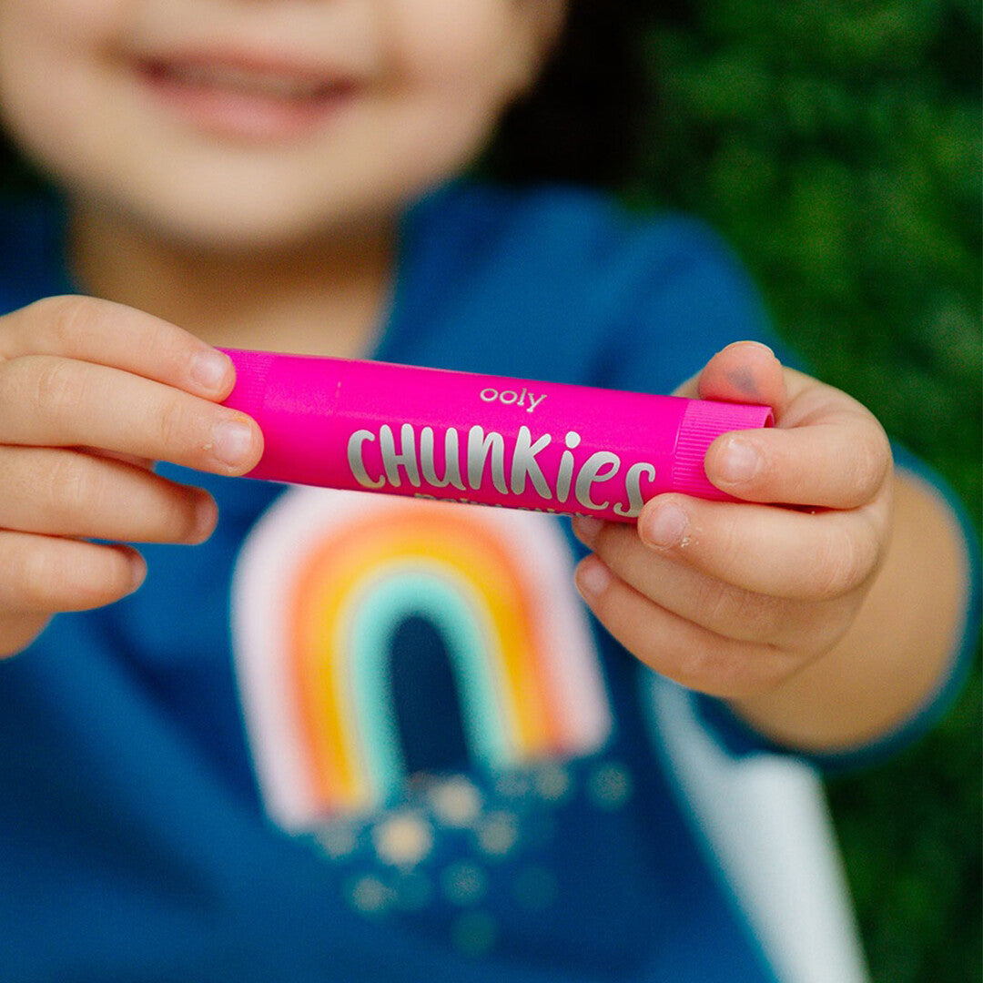 Kinderhanden houden een roze ooly Chunkies verfkrijtje vast, met een regenboog op de achtergrond.