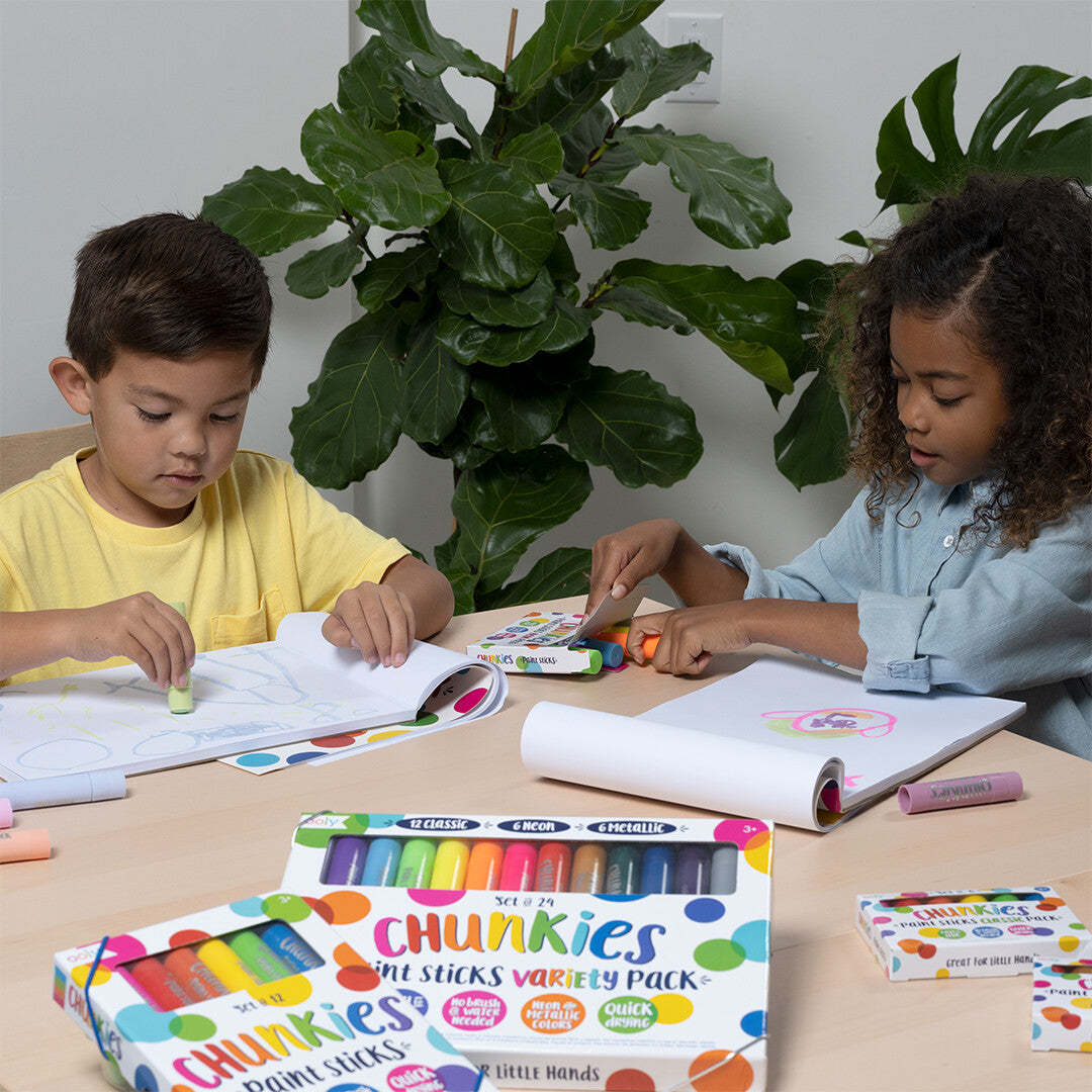 Twee kinderen tekenen met ooly Chunkies verfkrijtjes aan een tafel, met dozen verfkrijtjes op de voorgrond.